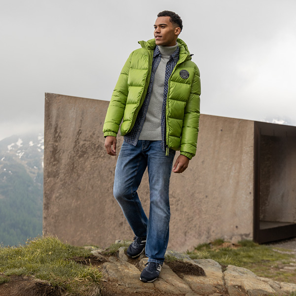 Mann in grünem, gepolstertem Jackett und Jeans steht auf einem Felsen mit Berglandschaft im Hintergrund.