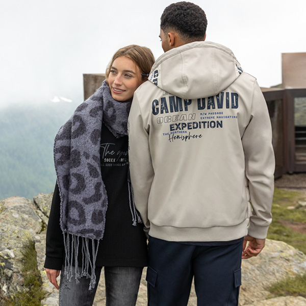 Paar in modischer Outdoor-Bekleidung, der Mann trägt einen grauen Hoodie, die Frau einen schwarzen Pullover mit Schal.