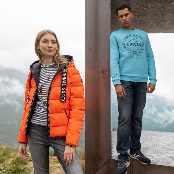 Zwei Modelle präsentieren modische Outfits: links eine Frau in einer orangefarbenen Jacke und gestreiftem Shirt, rechts e...