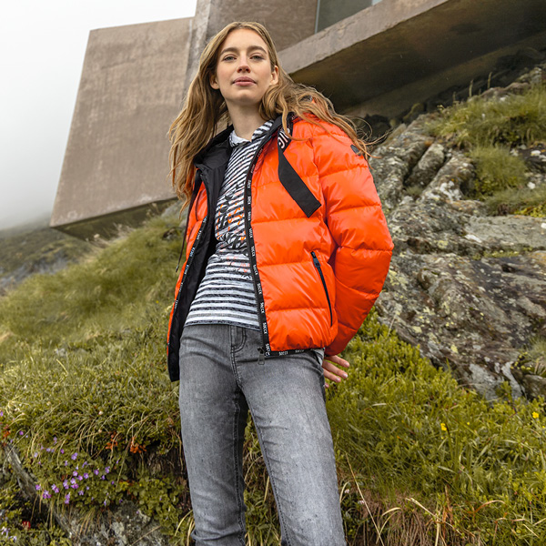 Junge Frau in orangefarbener Jacke und gestreiftem Shirt steht auf einer Wiese mit Felsen und modernem Gebäude im Hinterg...