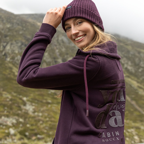 Frau in lila Hoodie und Mütze lächelt, vor einer bergigen Landschaft mit Wolken im Hintergrund.