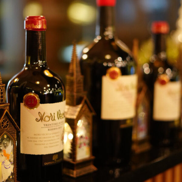 Dunkle Weinflaschen mit Etiketten stehen vor festlichen, dekorativen Häuschen auf einem Tisch.