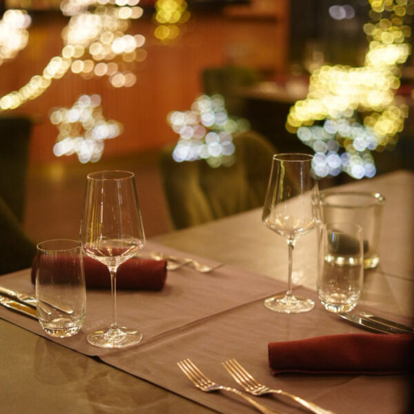Elegant gedeckter Tisch mit Gläsern und Besteck, im Hintergrund festliche Lichter und Weihnachtsdekoration.