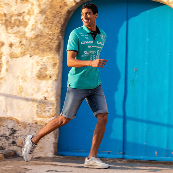 Junger Mann in türkisfarbenem Shirt und Jeansshorts läuft fröhlich vor einer blauen Tür.