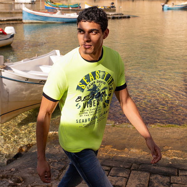 Junger Mann in leuchtend gelbem T-Shirt mit Grafikdesign, geht an einem Hafen mit Booten vorbei.