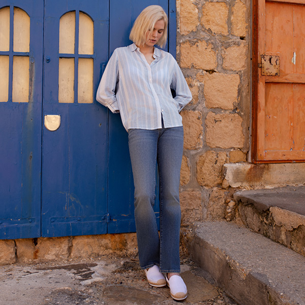 Junge Frau mit blonder, kurzer Frisur in gestreifter Bluse und Jeans steht vor einer blauen Tür aus Stein.