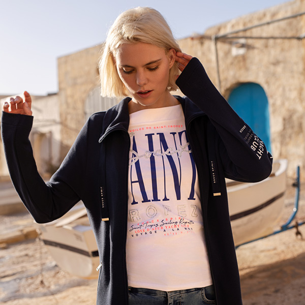 Frau mit blonden Haaren trägt ein weißes T-Shirt mit Aufdruck und eine dunkle Jacke, vor einem Boot in einer mediterrane...