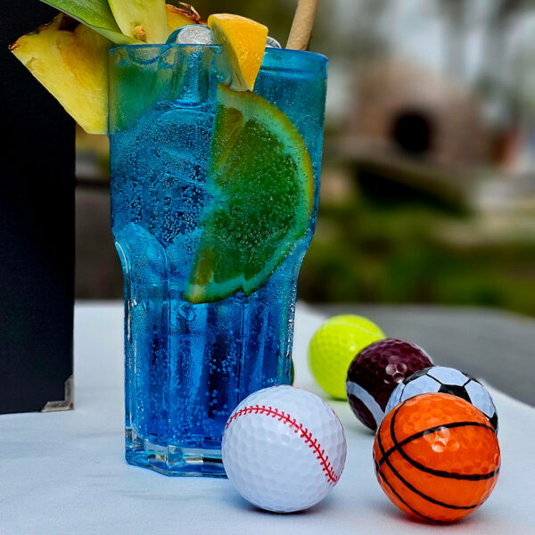 Blauer Cocktail mit Zitrusfrüchten in einem Glas, umgeben von bunten Miniatur-Sportbällen.