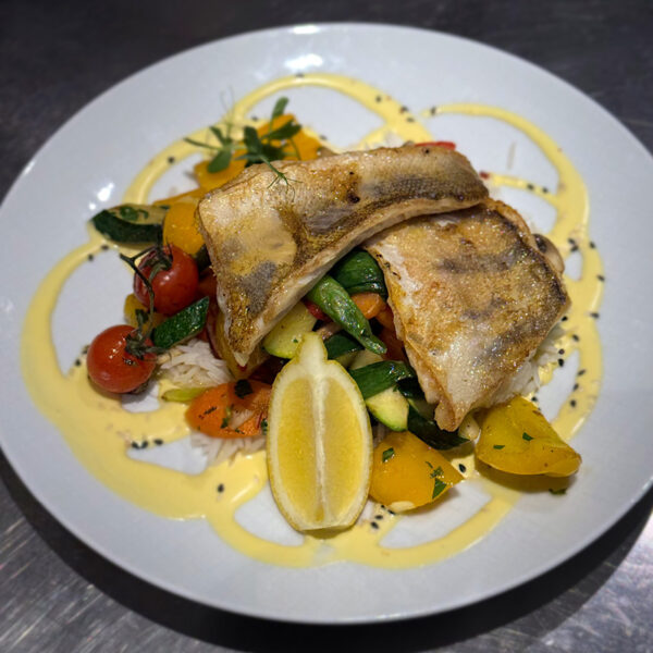 Gegrillter Fischfilet auf einem bunten Gemüsebett mit Zitronenscheibe und einer cremigen Sauce.