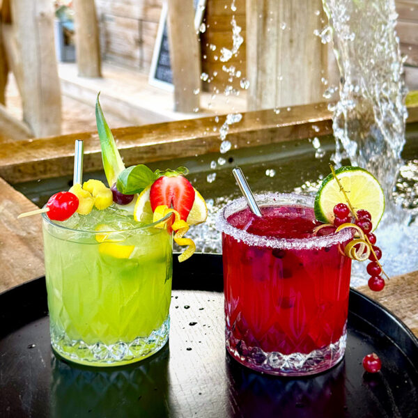 Zwei farbenfrohe Cocktails in Gläsern mit dekorativen Früchten, serviert auf einem Tablett vor einem Wasserfall.