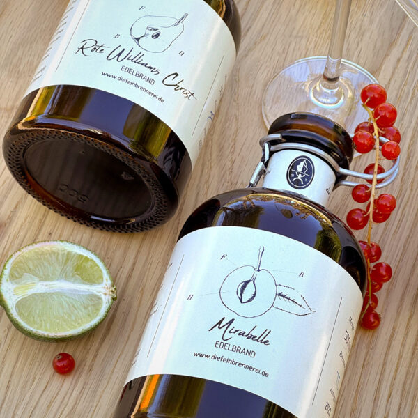 Zwei Flaschen Edelbrand mit Etiketten, eine mit Mirabelle und die andere mit Roter Williams, auf einem Holzuntergrund.