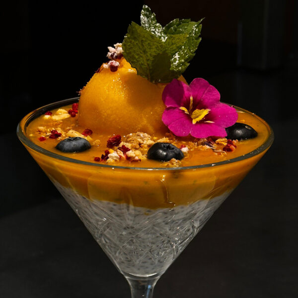 Fruchtiger Dessertbecher mit Mango-Sorbet, Beeren, Nüssen und einer essbaren Blüte, dekoriert mit Minze.