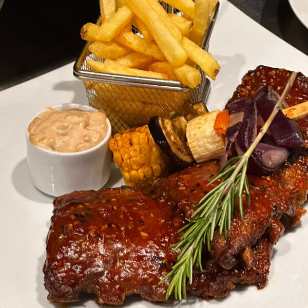 Saftige Rippchen mit BBQ-Sauce, Pommes frites, Maiskolben und einer cremigen Sauce auf einem weißen Teller.