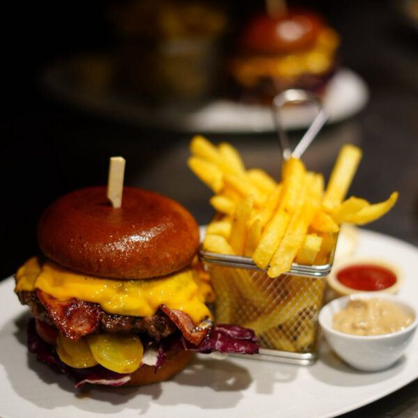 Saftiger Cheeseburger mit Bacon, Gurken und Käse, serviert mit Pommes frites in einem kleinen Korb und Dips.