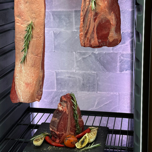 Drei Fleischstücke mit Rosmarin in einem Kühlschrank, darunter Gemüse auf einem schwarzen Stein.