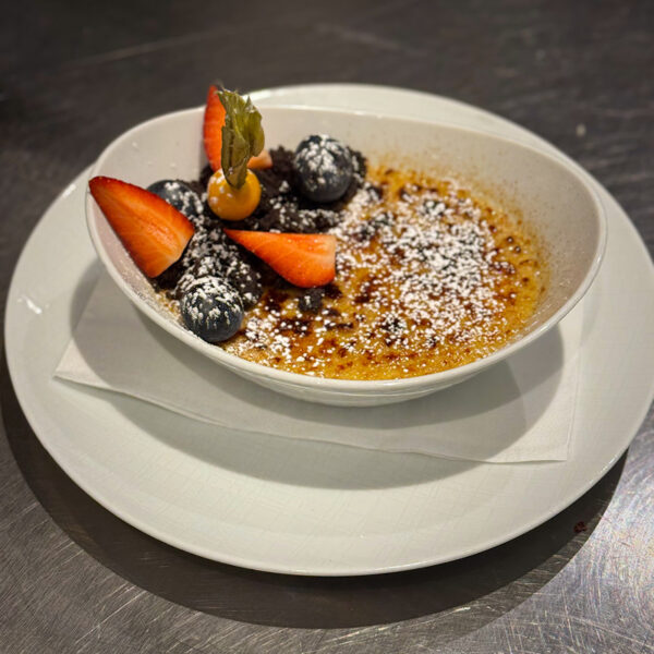 Creme Brûlée mit frischen Erdbeeren, Heidelbeeren und einer Physalis, serviert in einer weißen Schale auf einem Teller.